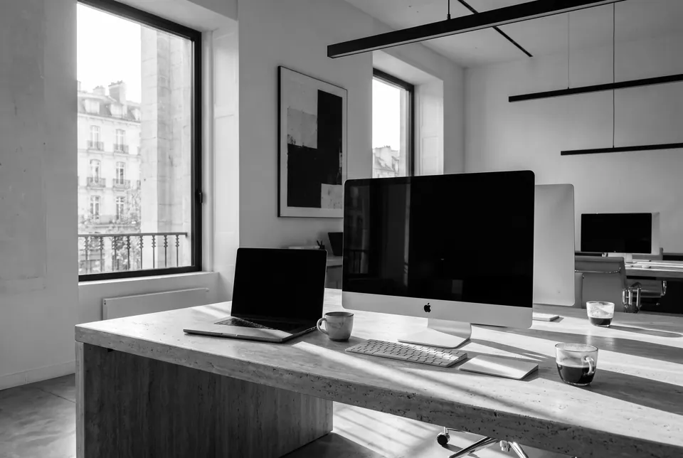 A desk by a tall window, with a desktop computer, a laptop and two cups of coffee in the sun