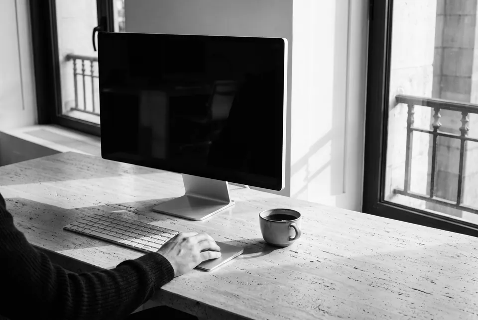 A hand on a keyboard at a stone desk, a monitor and a cup of coffee beside an open window